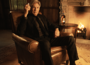 Harrison Ford raucht eine Zigarette und trinkt Whiskey neben einem Kamin.