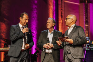 Drei Männer im Anzug stehen auf einer Bühne.