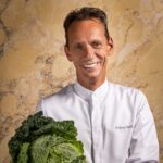 Ein Koch hält einen Kohl und lächelt in die Kamera.