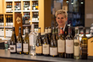 Frau steht hinter Theke mit Weinflaschen und Lächelt in die Kamera.