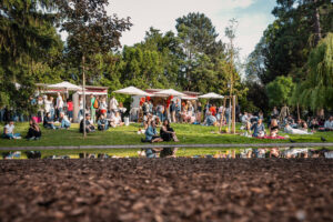 Leute sitzen in einem Park. Die Sonne scheint, es sind Zelte aufgebaut.