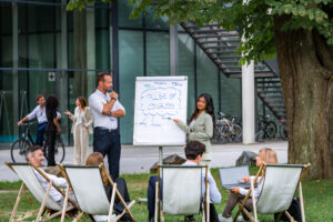 Eine Gruppe an Menschen hält im Freien einen Vortrag. Eine Frau und ein Mann stehen an einem weißen Flipchart.