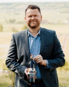 Ein Mann hält eine Weinflasche und lächelt in die Kamera. Er ist draußen und hat einen Anzug an.