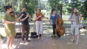 Eine Band spielt Musik auf einer Holzbühne. Im Hintergrund ist ein Wald zu sehen.