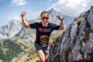 Ein Sportler mit Sonnenbrille und buntem T-Shirt zeigt seine Muskeln beim Lauf. Im Hintergrund sieht man die Alpen.