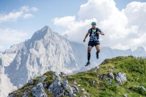 Ein Mann in Sportgewand springt über einen Stein. Im Hintergrund sieht man eine Berglandschaft.