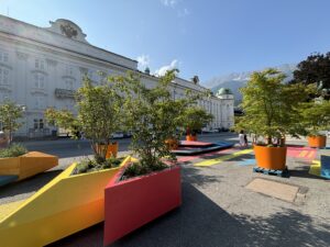 Grüne Bäumchen in bunten Trögen zieren einen Platz in Innsbruck. Im Hintergrund die Berge.